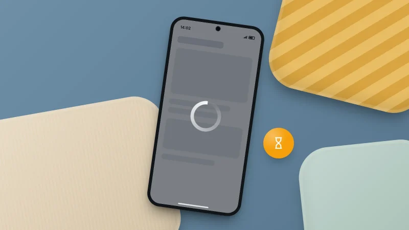 A phone lying among cushions, its dimmed screen frozen behind a loading circle, with an hourglass badge beside it