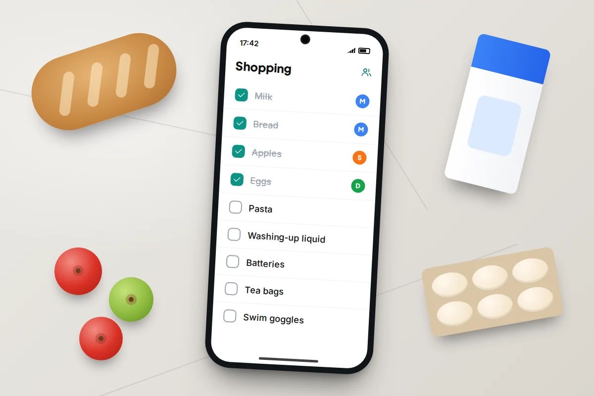 A kitchen worktop with bread, milk, eggs and apples around a phone showing a shopping checklist with items ticked