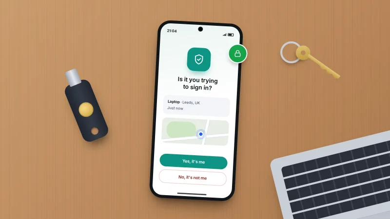 Phone on a wooden desk showing a sign-in approval prompt, beside a physical security key, a house key and a laptop